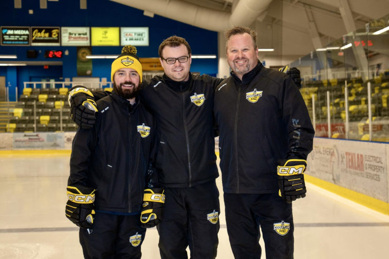 Three Coquitlam HC coaches in matching team jackets posing on the ice.