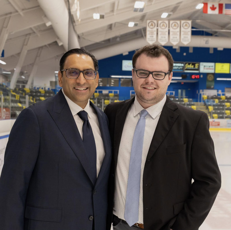 Two men in suits smiling in front of the ice with Canadian and U.S. flags hanging above the arena.