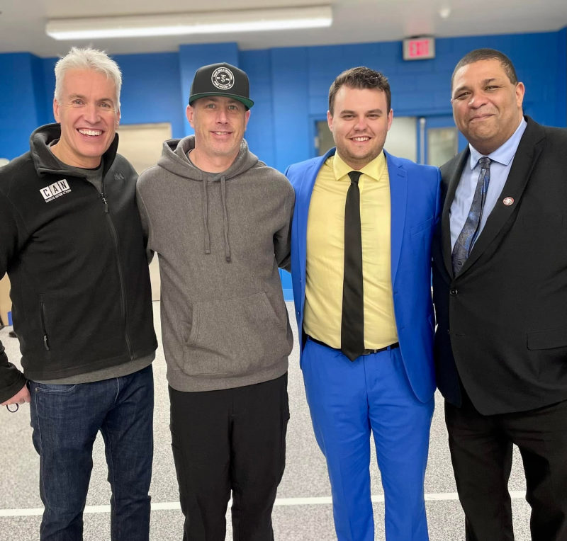 Four men posing together, including a representative in a Canucks Autism Network jacket at a community gym.