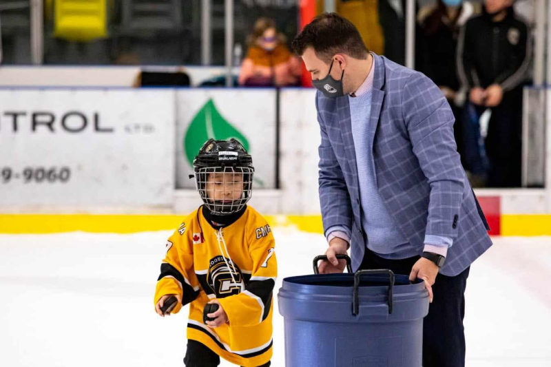 Tali Campbell in a plaid blazer collecting pucks with a young player in a yellow hockey jersey.
