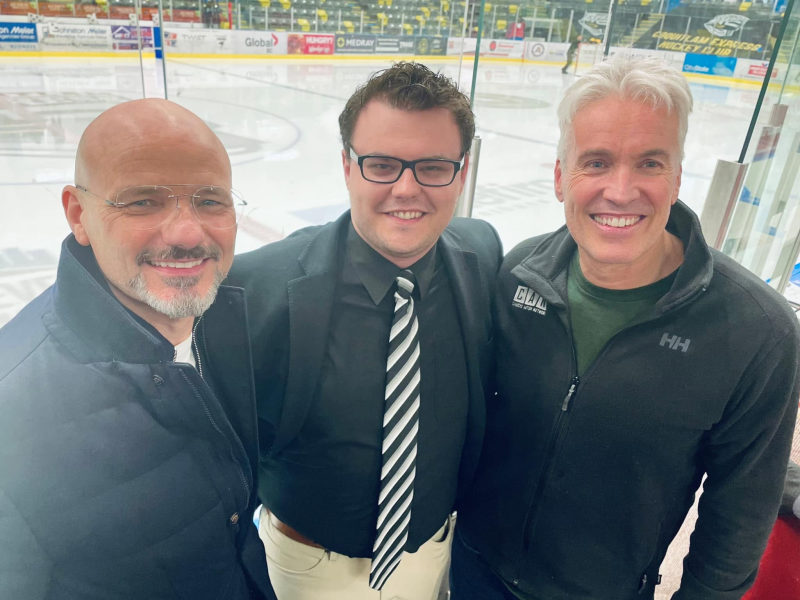 Three men smiling together in front of the Coquitlam Express rink boards and advertisements.