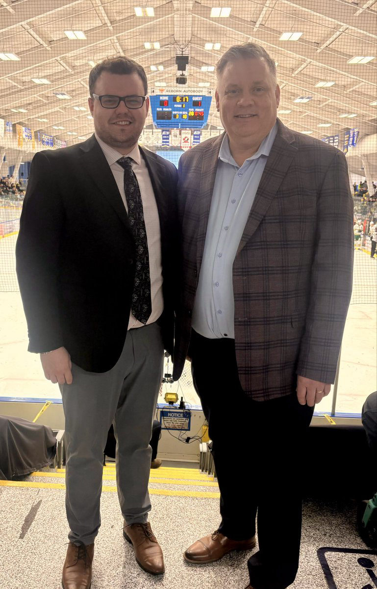 Tali Campbell in a black blazer smiling with a community partner in front of the arena netting and scoreboard.